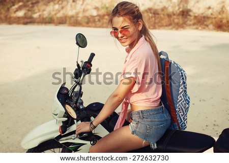 Outdoor summer portrait of young pretty blonde woman sitting on cross motorcycle traveling and having fun smiling with backpack