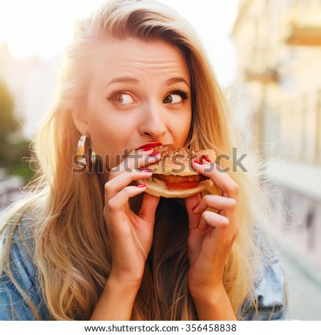 Outdoor fashion funny hipster style portrait of beautiful blonde woman eating tasty hamburger on the street