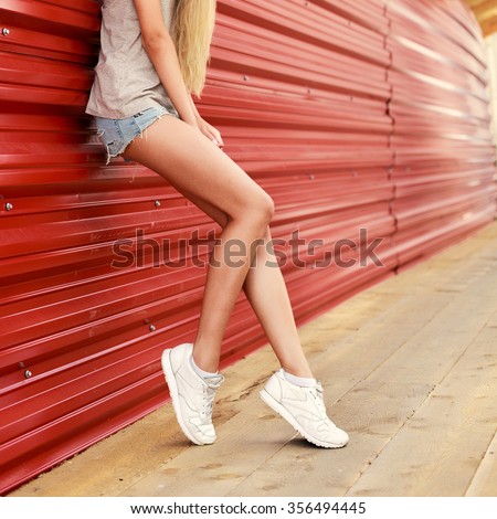 Beautiful young blonde long legs woman posing outdoor in summer sensual fashion hipster sport style on red background in jeans shorts and white sneakers