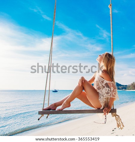 Summer portrait of beautiful stunning blonde woman sitting on swing on the beach on blue sea and green palms background smiling and having fun
