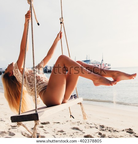Outdoor summer vacation tropic palm style portrait of young smiling blonde sexy woman posing in bikini on beach swing on sunset