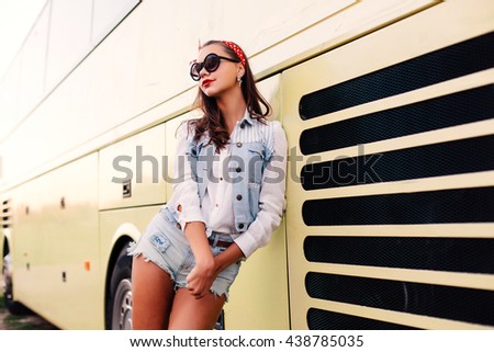 Pretty young girl posing near yellow bus in jeans shorts happy smiling in sunglasses with red lips having fun alone