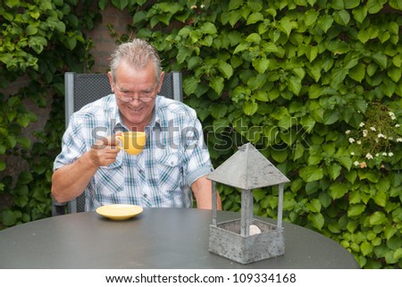 Retired senior Dutch man with a smile on his face whilst drinking a cup of hot coffee in his back yard