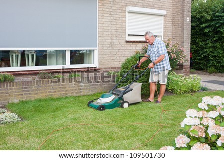 Dutch senior with serious look and mowing his front yard grass with an electric mower as spare time activity after retirement