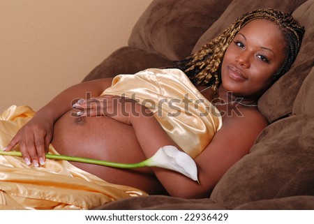 Beautiful pregnant African American woman, laying on a sofa wrapped in a gold satin sheet, she is holding a lily.