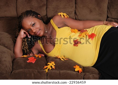 Beautiful pregnant African American woman, laying on the sofa wearing a yellow top. She has autumn leaves scattered on her because her twins are going to be born in autumn.