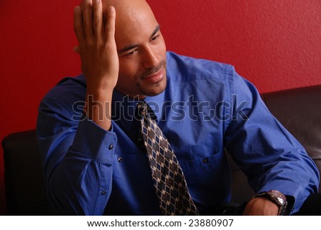 Attractive mixed race business man wearing a blue shirt and tie sitting on a leather sofa.  He is resting his head in his hand like he is thinking about something.