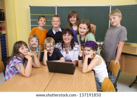 Teacher and children in a yellow classroom. They all looking to camera. The children are around the teacher, she is sitting in the middle. In front of the people you see a notebook.