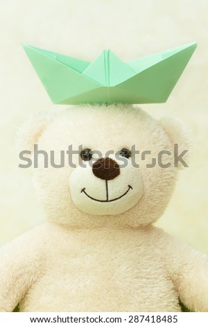 White teddy bear with a paper boat on a head.