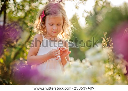 Cute blonde child girl in white dress plays in summer blooming garden