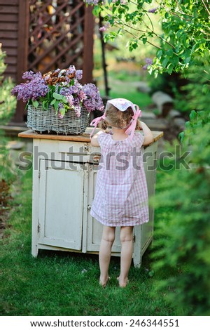 child girl in pink plaid dress standing near white vintage bureau with basket of lilacs in spring garden