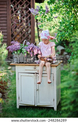cute little girl in pink dress sitting on vintage bureau and making lilac wreath in spring garden