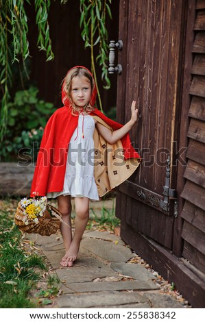 child girl playing little red riding hood in summer garden near wooden house