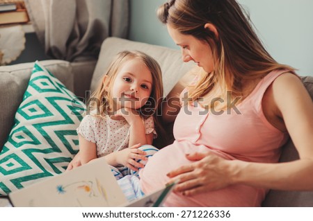 young pregnant mother reading book to her toddler daughter at home