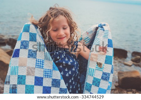 happy child girl at sea wrapped in quilt blanket, warm cozy mood on summer vacation