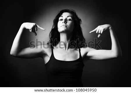 Strong woman demonstrating her muscles and pointing to herself. Black and white studio portrait