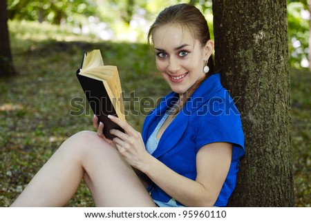 The beautiful girl the teenager in a dark blue blouse reads the book in park in the summer, having leaned against a tree and smiles