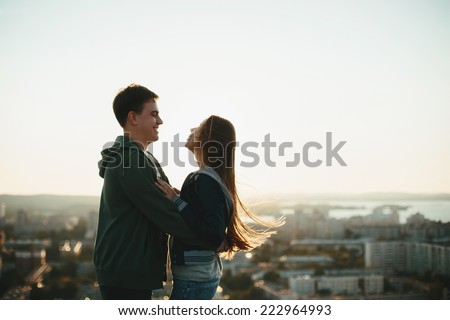 Sunny outdoor portrait of young happy stylish couple hugging on the roof at sunset. Man and woman laughing.