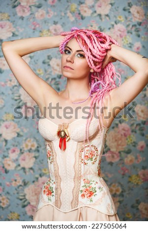 Freaky young female model in vintage clothing wearing corset with rose flower decoration against flower background. Bright pink dreadlocks hairstyle, beauty make up.