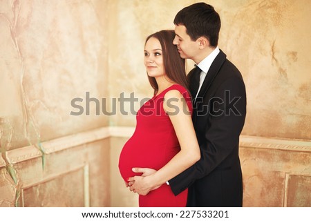 Stylish elegant young couple waiting a baby. Brunette female in red cocktail dress, man in tuxedo. Man hugging woman.