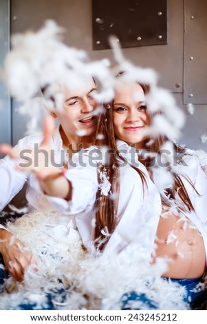 Young happy pregnant family have fun with fluffy feathers from pillow. Man and woman dressed in jeans and white shirts.