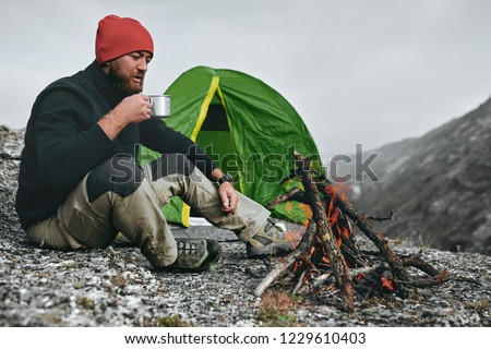 Outdoor shot of young breaded male drinking hot beverage in mountains. Traveler man in red hat, sitting near to camping tent, holding in hands a mug of tea after hiking. Travel, lifestyle.
