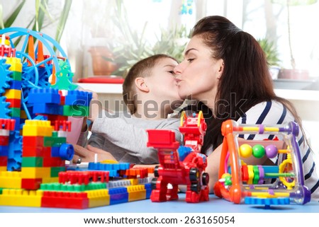 mother kissing her son sitting at a table with toys in home interior