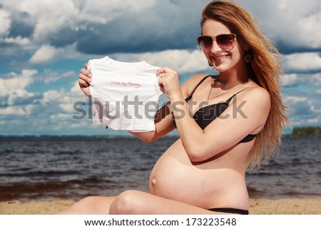 Pregnant sitting on the beach with little T-shirt pregnant holding a small tee shirt