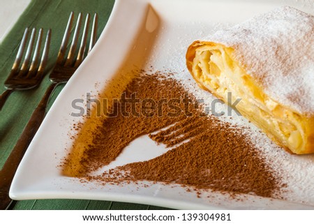 delicious strudel stuffed with cottage cheese, assign with powdered sugar on a white plate with a fork pattern of cocoa powder.