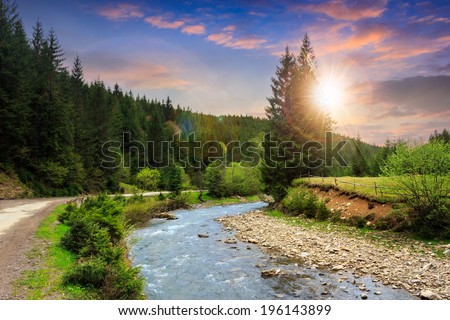 asphalt winding  road going along the river and passes through the green shaded forest in mountains at sunset