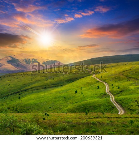 winding road through a large meadow on the hillside with some trees and far away mountain peak at sunset