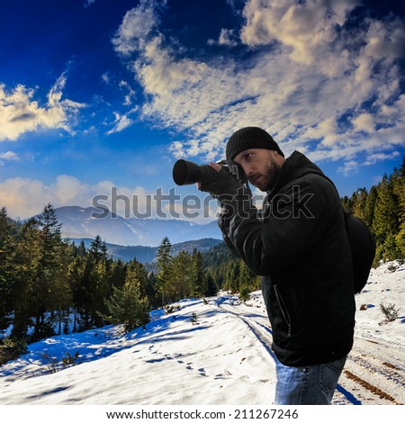 young photographer with camera and bag stands on snowy road to coniferous forest in mountains on sunrise