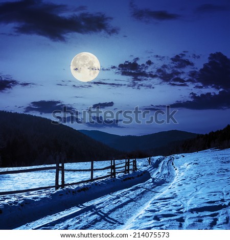 winter mountain landscape. winding road that leads into the pine forest covered with snow. wooden fence stands near the road at night in full moon light