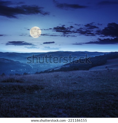 Stack of hay on a green meadow in the mountains in the morning under a blue summer sky at night in full moon light