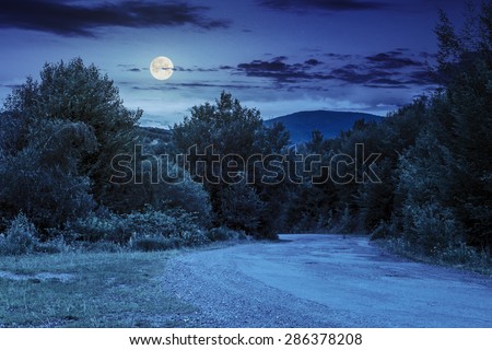 asphalt road going in mountains and passes through the green forest in shade of clouds at night in full moon light