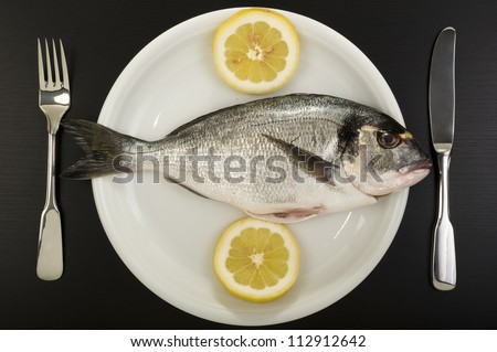 Fresh bream on a plate. Fresh sea bream on a plate with two pieces of lemon and silverware in black background. Studio shot