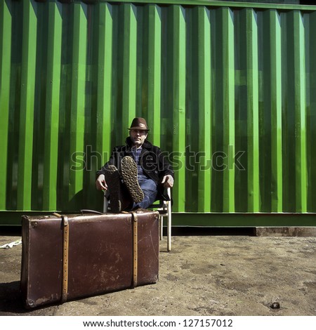Man sitting on a chair in a dockyard. A man with a suitcase is sitting on a chair against a container