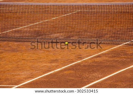 an empty tennis court with net and yellow ball close to it. It is a clear sunny day and the court is ready before to start a new game.