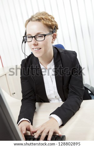 Attractive smart young secretary or corporate personal assistant wearing a headset sitting at her desk typing on her computer