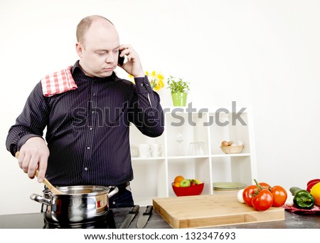 Attractive middle-aged balding man taking a call on his mobile while standing cooking at the stove stirring the contents of a stainless steel saucepan