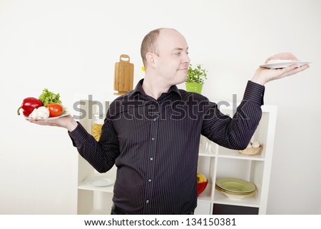 Gleeful smiling man standing in his kitchen with his arms raised with two plates, one with fresh vegetables and one with meat, making a choice on food and diet and giving in to temptation