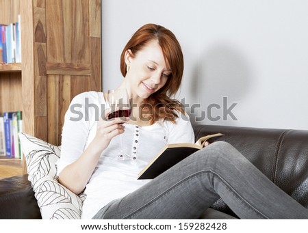 Pretty young redhead woman reading a book reclining on a black leather sofa with a glass or red wine in her hand and a smile of contentment on her face