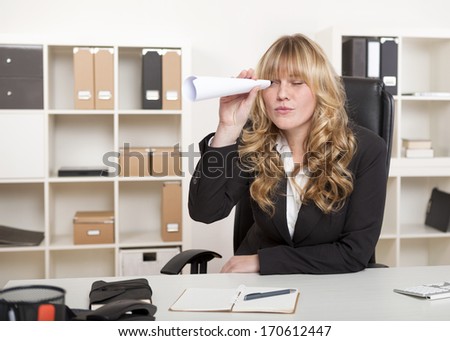 Bored young businesswoman sitting at her desk in the office playing with a paper telescope made from a rolled sheet of paper holding it to her eye