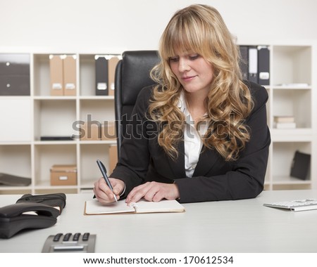 Blond businesswoman writing notes at her desk as she sits in her office with shelving lined with binders behind her