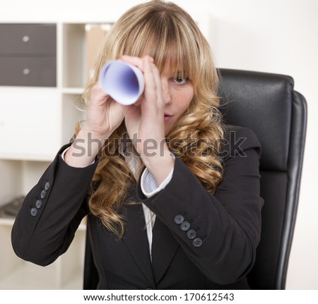 Businesswoman playing - I Spy - with a rolled up sheet of paper pretending it is a telescope as she sits at her desk in the office