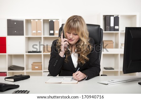 Attractive blond businesswoman or manageress chatting on the phone in the office as she sits at her desk taking notes as she listens to the conversation