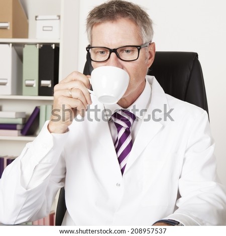 Middle-aged male doctor drinking a cup of tea or coffee as he sits at his desk, in his office looking down at his desk with downcast eyes.