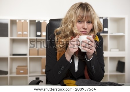 Cold businesswoman warming up with a mug of hot coffee as she sits at her desk, in the office wearing a knitted winter scarf.
