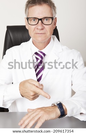 Serious Male Doctor Pointing his Wrist Watch, Looking at Camera