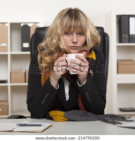 Young girl at work sitting at her desk in the office with a warm winter scarf around her neck enjoying a mug of hot coffee